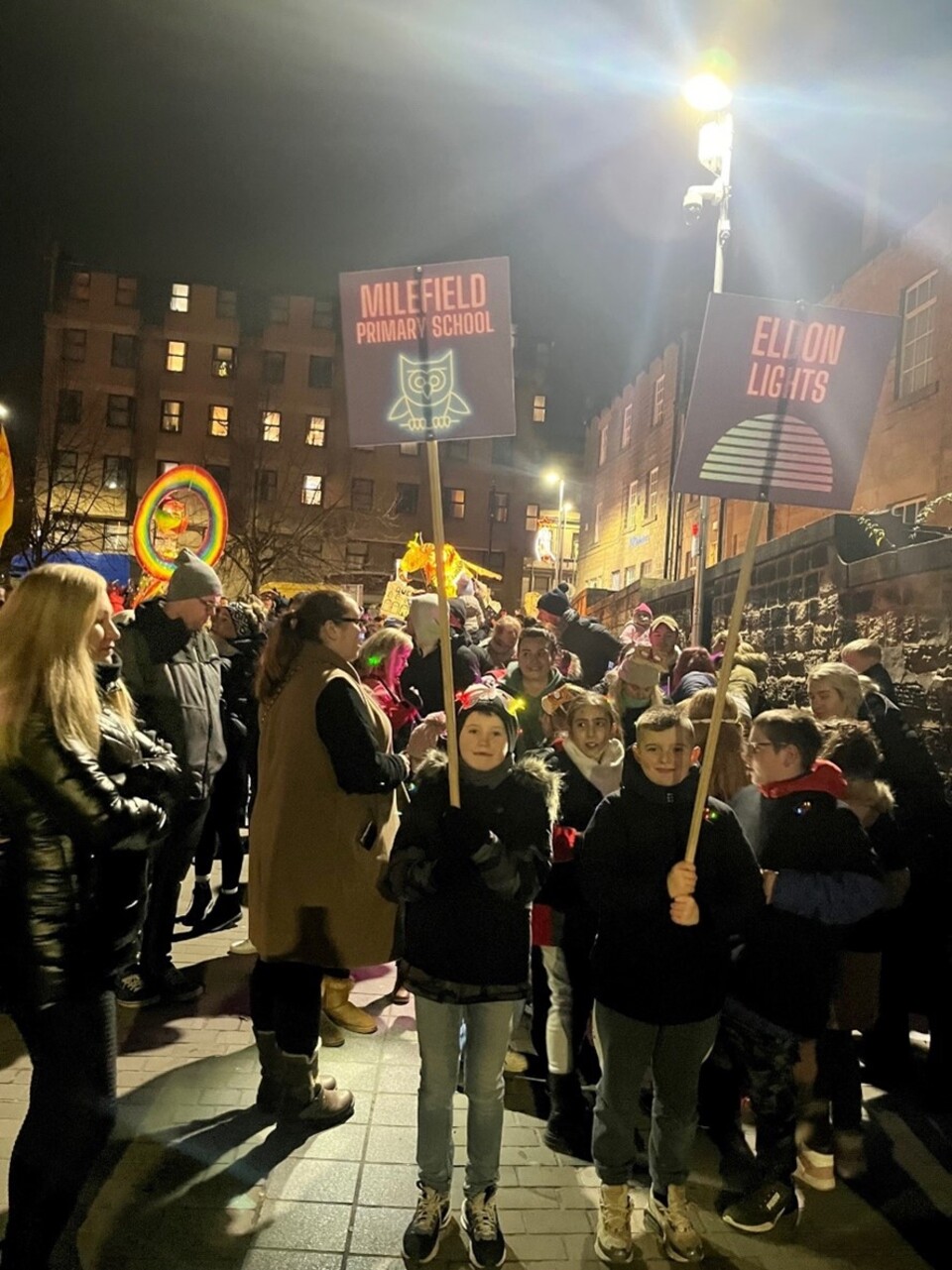 A group of children and adults stand outdoors at night during a festive event. Two children in front hold signs reading "Millfield Primary School" and "Eldon Lights." Bright lights and colourful decorations are visible.